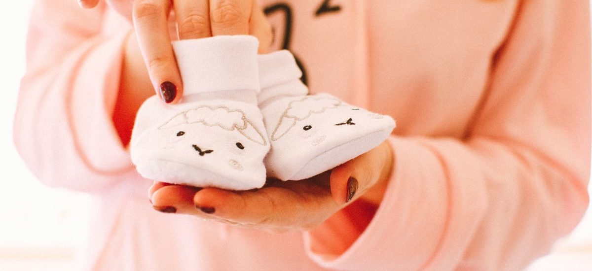 A woman in pink holds cute baby shoes with sheep designs, symbolizing warmth and love.